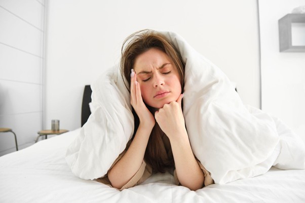 Woman lying in bed under a white duvet, pressing her temple in apparent headache pain.