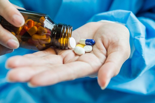 Medical professional pours pills from a brown bottle into an open palm, with assorted capsules and tablets visible in the hand and blue scrubs in the background.
