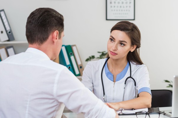 Female clinician with stethoscope consulting a male patient across a desk in a bright clinic room.