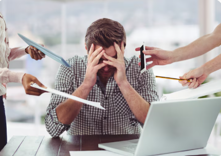 Man at a desk with a laptop looks overwhelmed as colleagues push papers and a phone toward him in a tense office scene.