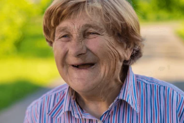 Smiling elderly woman with short light-brown hair wearing a striped shirt outdoors on a sunny day.