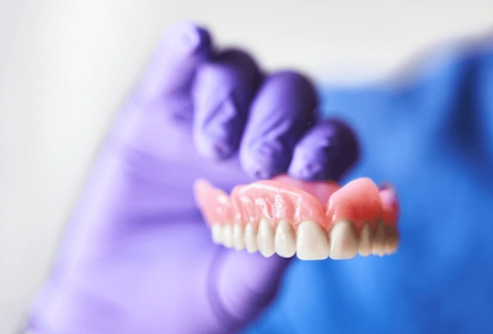 Dentures held by a gloved hand, showing pink gum-colored base and white teeth against a blue background.