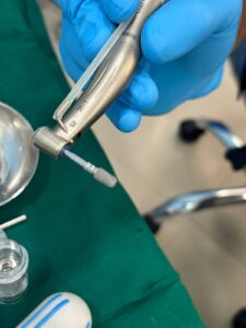 Close-up of a dentist's hand in blue gloves holding a metallic dental handpiece with a small attachment and suction tube in a clinical setting