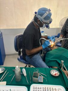 Dentist in a surgical headlamp and mask performing a dental procedure on a patient, with instruments laid out on a green tray nearby.