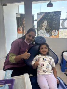 Dentist in maroon scrubs giving a thumbs-up beside a smiling young girl in a dental chair.
