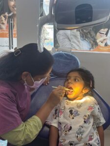 Dental clinician in pink scrubs and mask checks a child's mouth with a toothbrush inside a mobile dental clinic bus.
