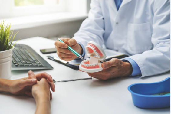 Dentist in a white coat demonstrates brushing technique with a dental model to a patient at a desk.