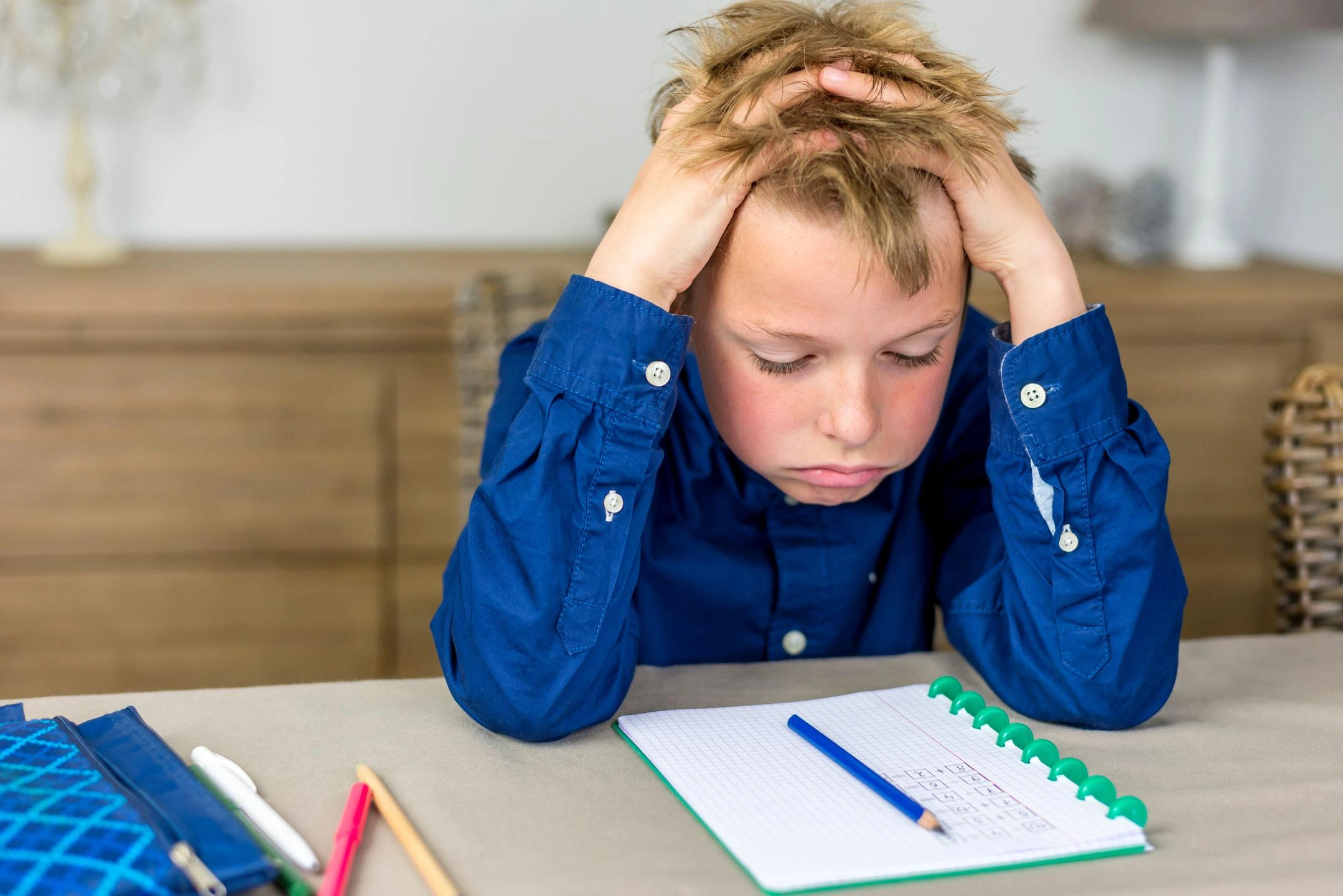 Boy with blond hair in a blue shirt holds his head in distress while doing homework at a table with a notebook and pencils nearby.