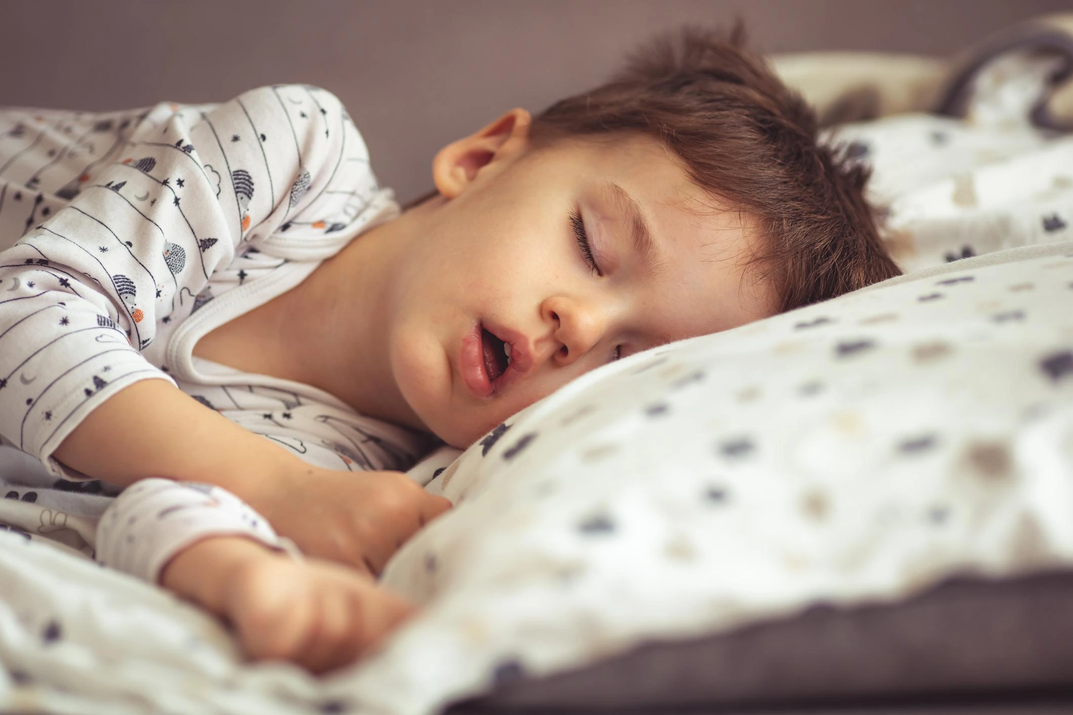 Young child asleep on a bed, lying on their side under patterned white sheets with a calm expression.