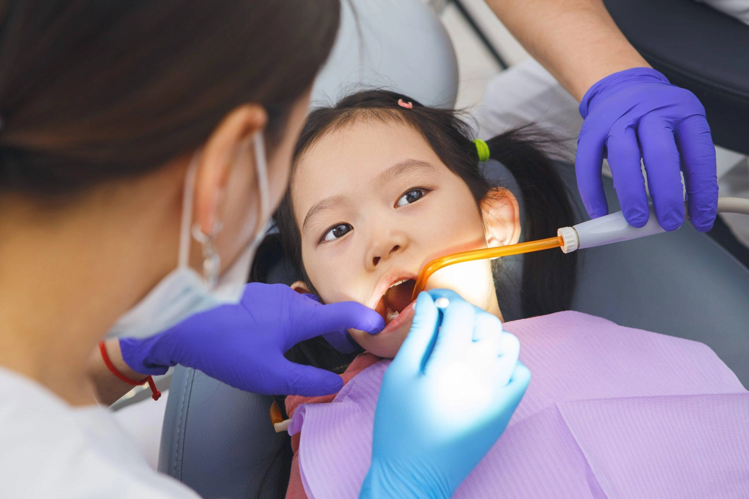 Pediatric dentist in purple gloves examining a young girl's teeth in a dental chair.